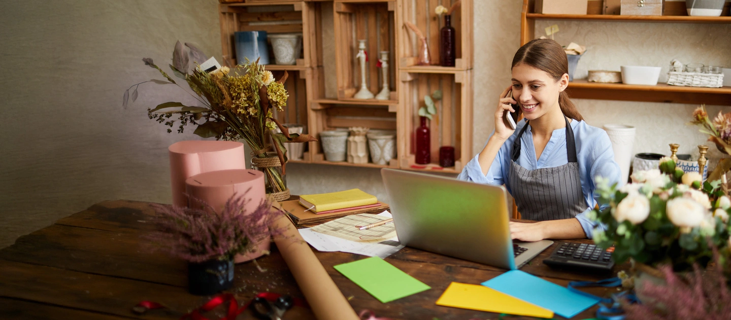 florist in their store sitting at their desk using a laptop and talking on the phone