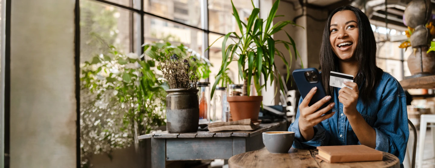 person sitting at a cafe table with a hot beverage holding their credit card and phone while smiling