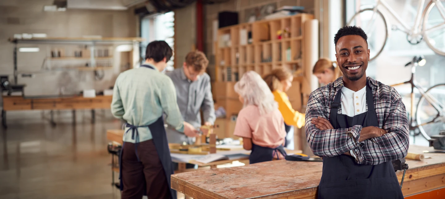 shop owner smiling and posing for the camera as a group of people partake in a workshop behind the owner