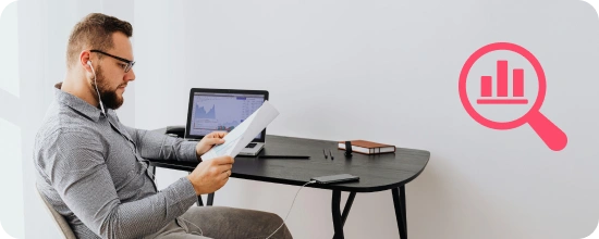person sitting at a desk with a laptop on it while they review information on paper and using earbuds to listen to their phone