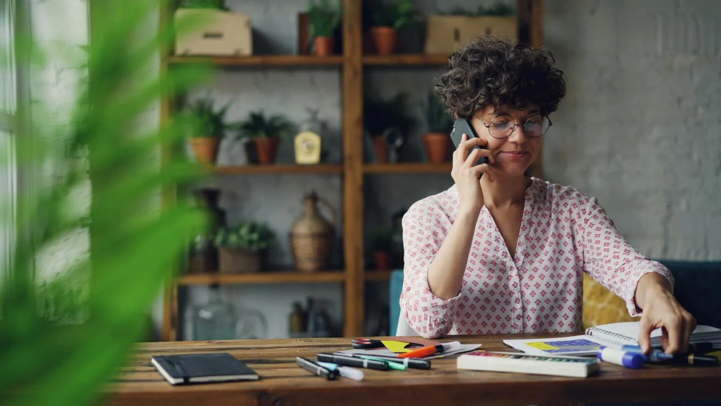person sitting at a table with artbook and art supplies talking on their mobile phone