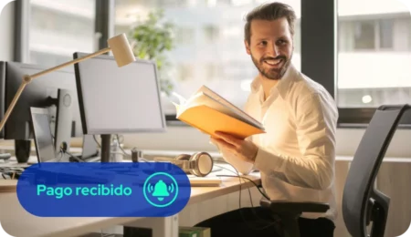 Persona sentada en su escritorio frente a un computador, sosteniendo un cuaderno y sonriendo